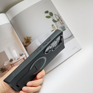 May include: A black phone case with a built-in stand and a circular magnetic attachment. The case has a sleek, matte finish and features buttons on the side for volume and power. The phone case is designed to protect the phone.