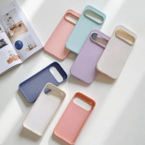 Puede incluir: Una colección de fundas para teléfonos de colores pastel, rosa, azul, morado y blanco. Las fundas son rectangulares con un recorte para la cámara y están dispuestas sobre una superficie blanca, lo que sugiere una estética moderna.