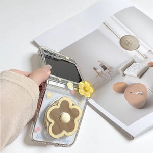 May include: Clear phone case with a brown and cream flower design and a yellow flower pop-up grip. The case has a floral pattern and the text "Have a nice day".