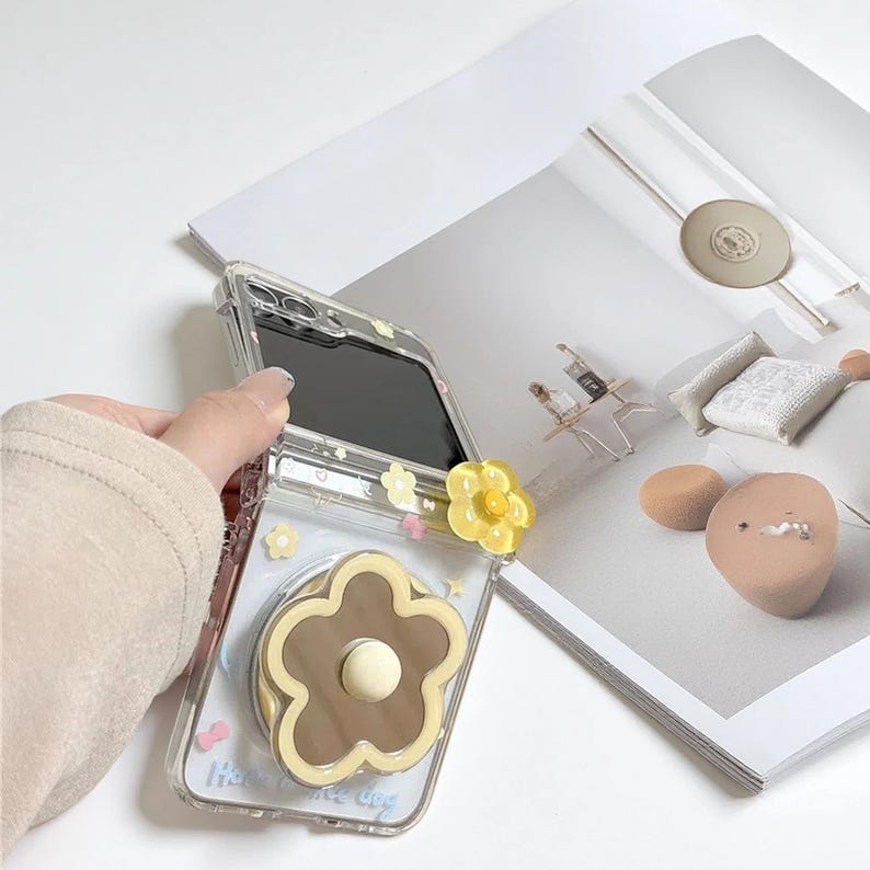 May include: Clear phone case with a brown and cream flower design and a yellow flower pop-up grip. The case has a floral pattern and the text "Have a nice day".