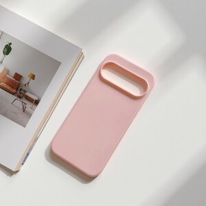 May include: A light pink phone case with a rectangular design and a raised camera lens protector. The case is made of a smooth material and is shown on a white surface next to an open magazine.