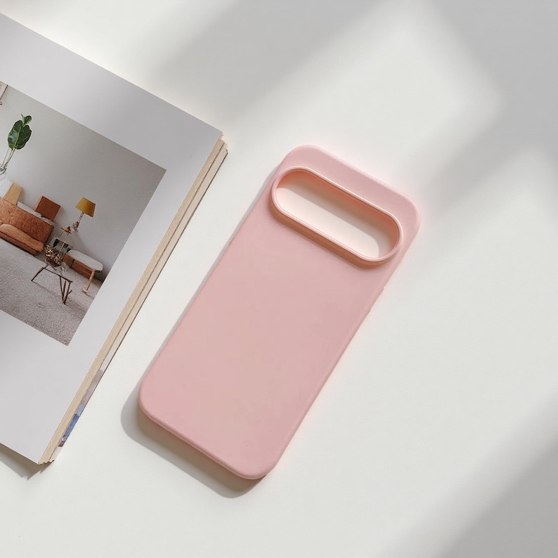 May include: A light pink phone case with a rectangular design and a raised camera lens protector. The case is made of a smooth material and is shown on a white surface next to an open magazine.