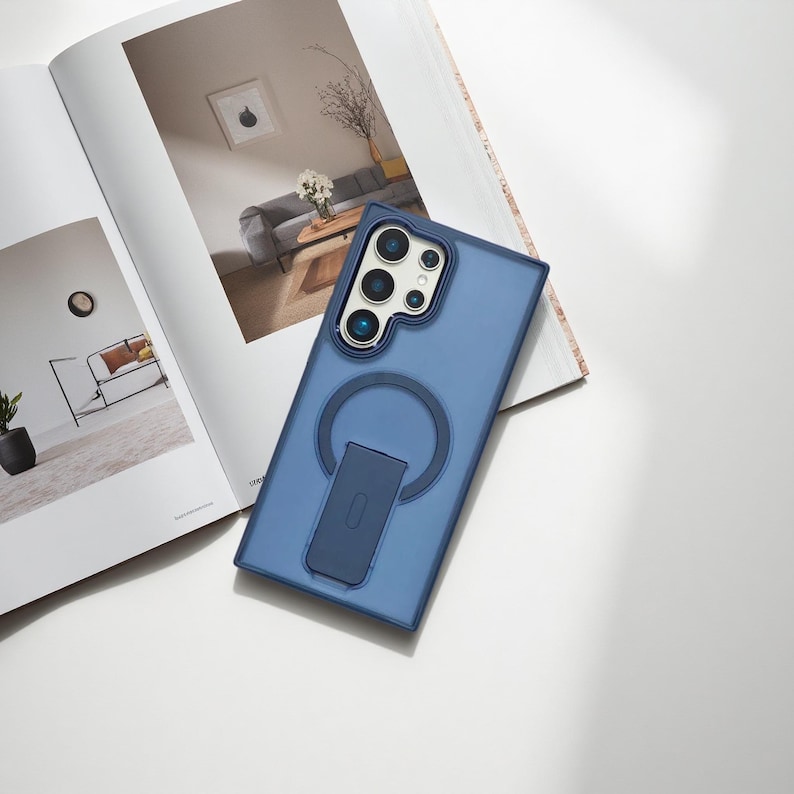 May include: A blue phone case with a circular magnetic ring and a kickstand. The case has a camera cutout with multiple lenses. The phone case is lying on top of an open magazine with interior design photos.
