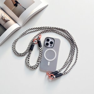 May include: A grey phone case with a white circular design and a black and white rope strap. The strap has black accents and orange metal rings. The phone case is lying on a white surface.