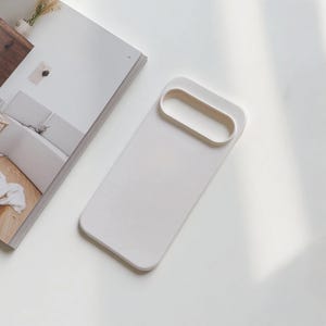 May include: A white rectangular phone case with a rounded rectangular cutout at the top. The case is lying on a white surface next to an open magazine.