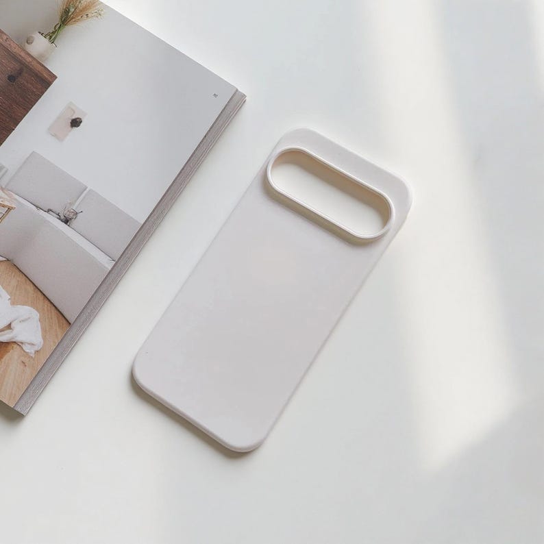 May include: A white rectangular phone case with a rounded rectangular cutout at the top. The case is lying on a white surface next to an open magazine.