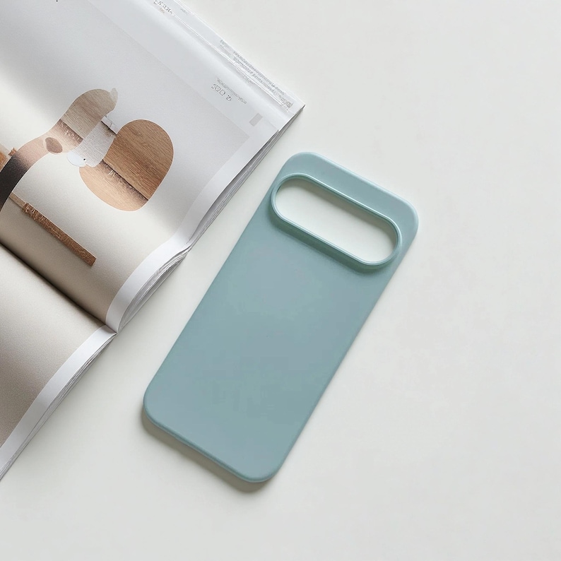 May include: A light blue phone case with an oval camera cutout. The case is rectangular with rounded edges. The case is lying on a white surface next to an open magazine.