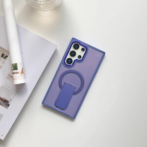 May include: A lavender phone case with a blue border and a matching kickstand. The case has a circular ring and a rectangular stand for hands-free viewing. The camera area has a blue border. The case is on a white surface.