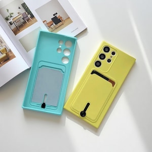 Puede incluir: Dos fundas para teléfono, una turquesa y otra amarilla, con ranuras para tarjetas. La funda amarilla tiene un módulo de cámara con múltiples lentes. Ambas fundas están sobre una superficie blanca junto a una revista abierta.