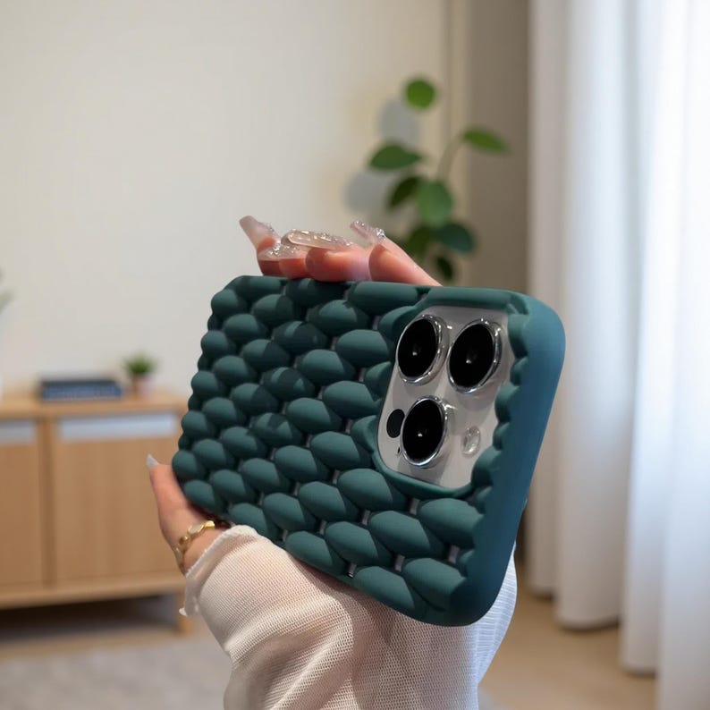 May include: Teal phone case with a textured, bubble-like pattern. The case has a smooth, teal border around the camera and phone edges. The camera lenses are visible. The case is held in a person's hand.
