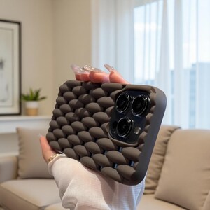 May include: A dark gray phone case with a textured, woven pattern. The case has a raised, bubble-like design and a dark gray border. The camera lenses are visible on the back of the phone. The case is designed to protect the phone.