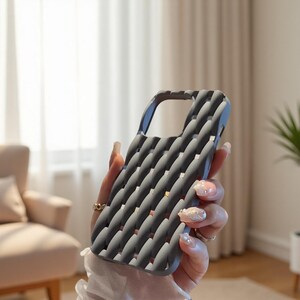 May include: A gray phone case with a textured, woven pattern. The case has a curved design with a cutout for the camera and buttons. The case is held in a person's hand, showcasing its design and texture.