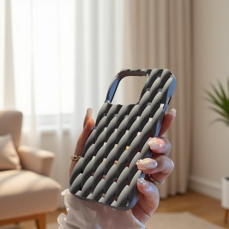 May include: A gray phone case with a textured, woven pattern. The case has a curved design with a cutout for the camera and buttons. The case is held in a person's hand, showcasing its design and texture.