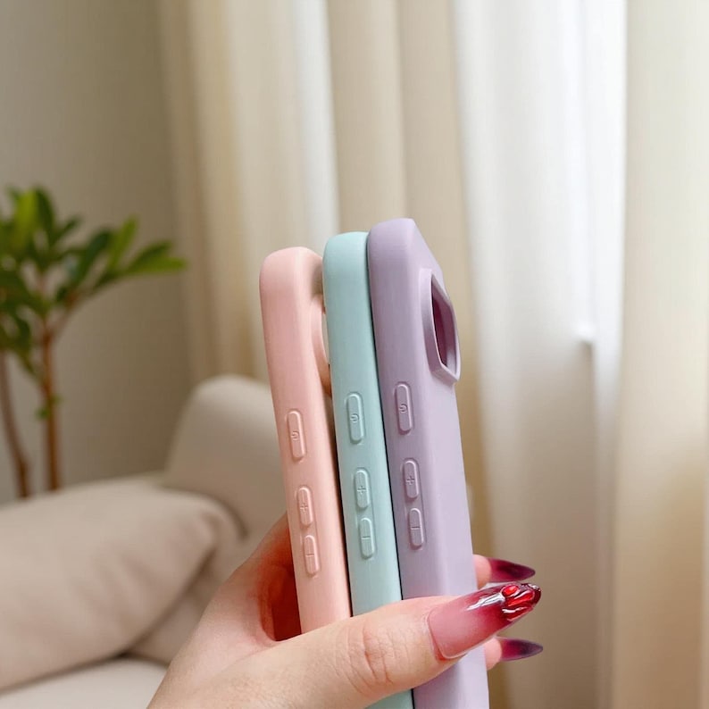 May include: Three pastel-colored phone cases in pink, mint green, and lavender. The cases are made of a smooth material and have button and port cutouts. The cases are held in a person's hand, with the focus on the phone cases.