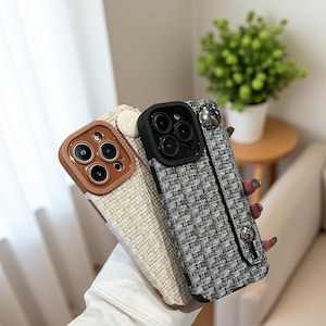 May include: Three phone cases in a close-up shot. One is brown with a leather effect, another is off-white, and the third is black and white tweed. Each case has a camera lens protector and a wrist strap.