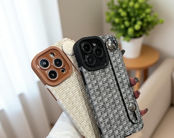 May include: Close-up of three phone cases. One case is brown with a leather texture, another is off-white, and the third is black and white tweed. Each case has a camera lens protector and a wrist strap.