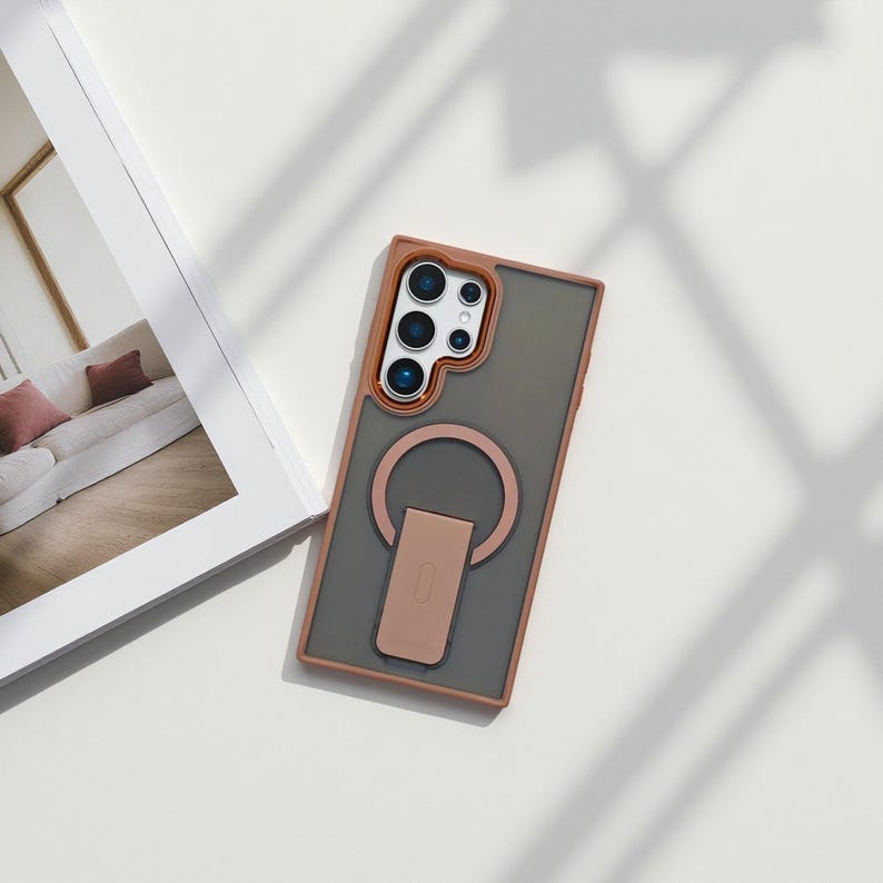 May include: A phone case with a gray back, brown frame, and a brown ring holder. The case has a camera cutout with multiple lenses. The ring holder is in the shape of a C and has a small rectangular stand.