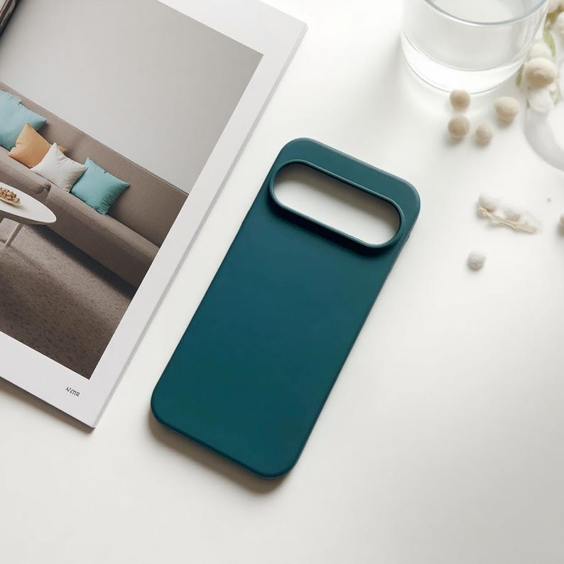 May include: Teal-colored phone case with a rectangular design and a large camera cutout. The case is made of a smooth material and is shown on a white surface next to a magazine.