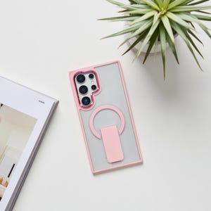 May include: A clear phone case with a pink frame and a pink ring holder. The case has a clear back and a pink border around the camera lenses. The ring holder is attached to the back of the case.