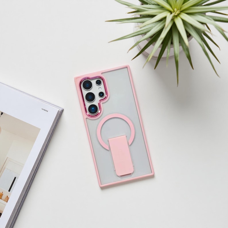 May include: A clear phone case with a pink frame and a pink ring holder. The case has a clear back and a pink border around the camera lenses. The ring holder is attached to the back of the case.