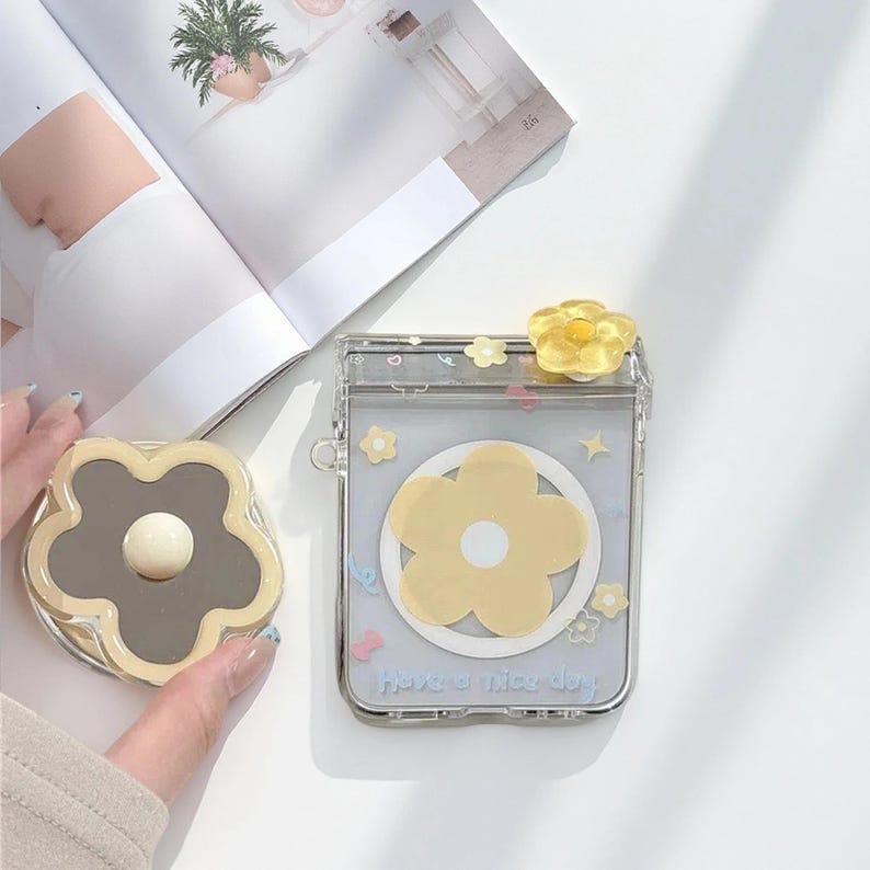 May include: Clear case with a yellow flower design and the text "Have a nice day." A matching compact mirror with a flower shape and a gray center is also visible. The case has a yellow flower on top.