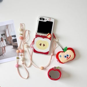 May include: A clear phone case featuring a red apple design, a matching apple-shaped plushie, and a small apple-shaped mirror. The case has a pink strap with decorative beads. The plushie has a smiling face and blue accents.