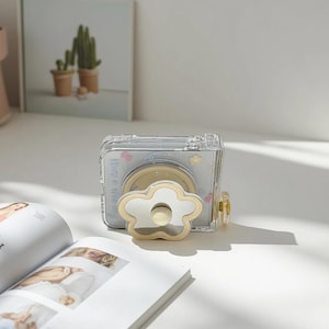 May include: Clear, rectangular phone case with a flower-shaped stand. The case has a light blue background with small flower and heart designs. The text "Have a nice day" is printed on the case. A magazine and a framed picture of cacti are in the background.