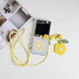 May include: Clear phone case with a yellow flower design and a matching yellow lanyard. The case has a yellow flower charm and a crocheted yellow flower with a green leaf. The phone screen is black. The case says "Have a nice day".