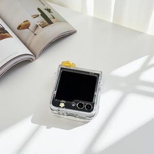 May include: Clear phone case with a black screen, featuring a yellow flower accent and a small floral design. The case has a transparent design, allowing the phone's features to be visible. The phone case is on a white surface next to an open magazine.