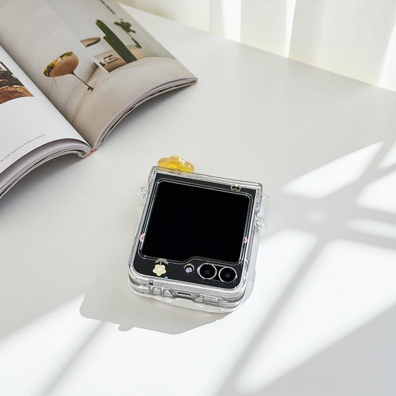 May include: Clear phone case with a black screen, featuring a yellow flower accent and a small floral design. The case has a transparent design, allowing the phone's features to be visible. The phone case is on a white surface next to an open magazine.