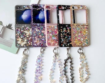 May include: A collection of bedazzled phone cases in various colors, including silver, purple, pink, black, and gold. Each case features a butterfly or abstract design with sparkling rhinestones. Attached are beaded wrist straps.