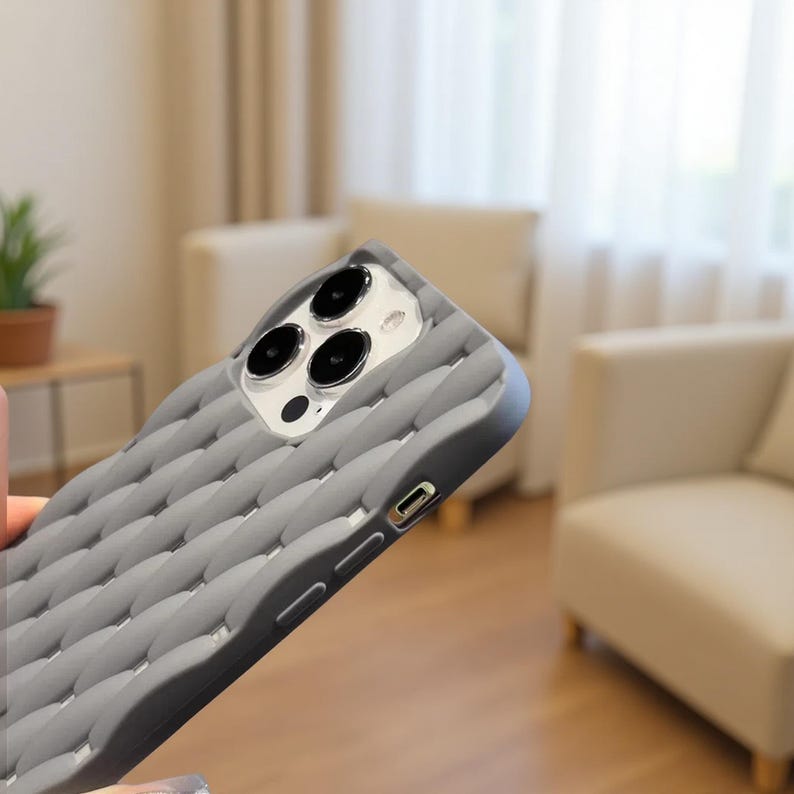 May include: A gray phone case with a textured, woven pattern. The case has a dark gray border and cutouts for the camera lenses and charging port. The phone case is designed to protect the phone.