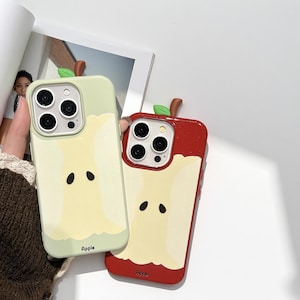 May include: Two phone cases, one light green and one red, each designed to resemble a sliced apple. The cases feature a green leaf and brown stem detail at the top. The apple design includes a yellow interior with black seed details and the word "Apple" at the bottom.