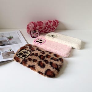 Op de afbeelding: Een verzameling pluizige telefoonhoesjes in verschillende kleuren en patronen. De hoesjes omvatten een bruin-zwarte luipaardprint, een roze hoesje, een wit hoesje en een roze-rode luipaardprint. Elk hoesje heeft een cameralensbeschermer.