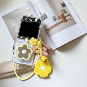 May include: Clear phone case with a daisy design and a yellow strap. The case has a large gray flower with a white center and a smaller yellow flower charm. A yellow crocheted flower with a bell is attached to the strap.