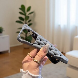 May include: A gray phone case with a textured, geometric pattern. The case has cutouts for the camera and buttons. The case is held in a hand with gold rings. The background is blurred, showing a plant and a white wall.