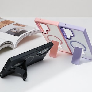 May include: Three phone cases in the image. One black case with a built-in stand, and two clear cases with colored frames in pink and purple. The cases have a circular design on the back and are designed to stand upright.