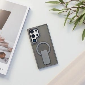 May include: A gray phone case with a built-in ring holder and camera cutouts. The case has a rectangular design with a raised edge for protection. The ring holder is silver and can be used as a stand. The phone case is on a white surface.