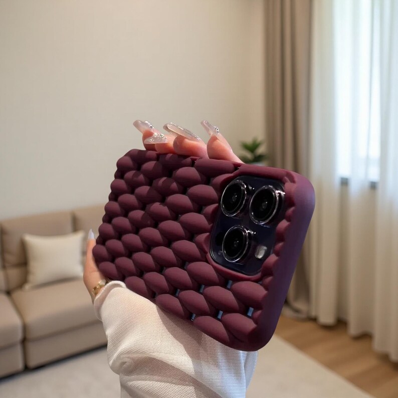 May include: A burgundy phone case with a textured, bubble-like pattern. The case has a raised edge around the camera lenses. The phone case is held in a person's hand.