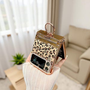 May include: A gold-toned phone case featuring a leopard print pattern. The case includes a ring holder and is adorned with rhinestones around the camera and screen. The phone screen shows the time as 12:45.