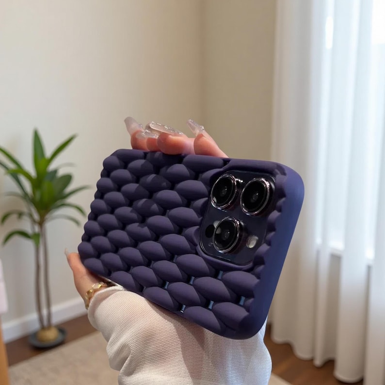 May include: A dark purple phone case with a textured, bubble-like pattern. The case has a raised edge around the camera lenses and a smooth, matte finish. The case is designed to protect the phone from scratches and impacts.