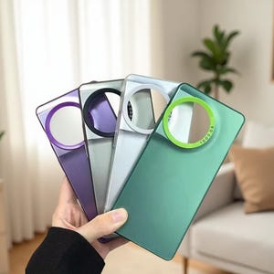 May include: A hand holding four phone cases in shades of purple, gray, white, and green. Each case has a circular design element with the text "SO COOL" around the edge. The cases have a sleek, modern design.