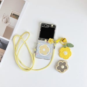 May include: A clear phone case with a yellow flower design and a matching yellow lanyard. The case is attached to a yellow crocheted flower charm with a green leaf and a small bell. A second flower-shaped accessory is also present.