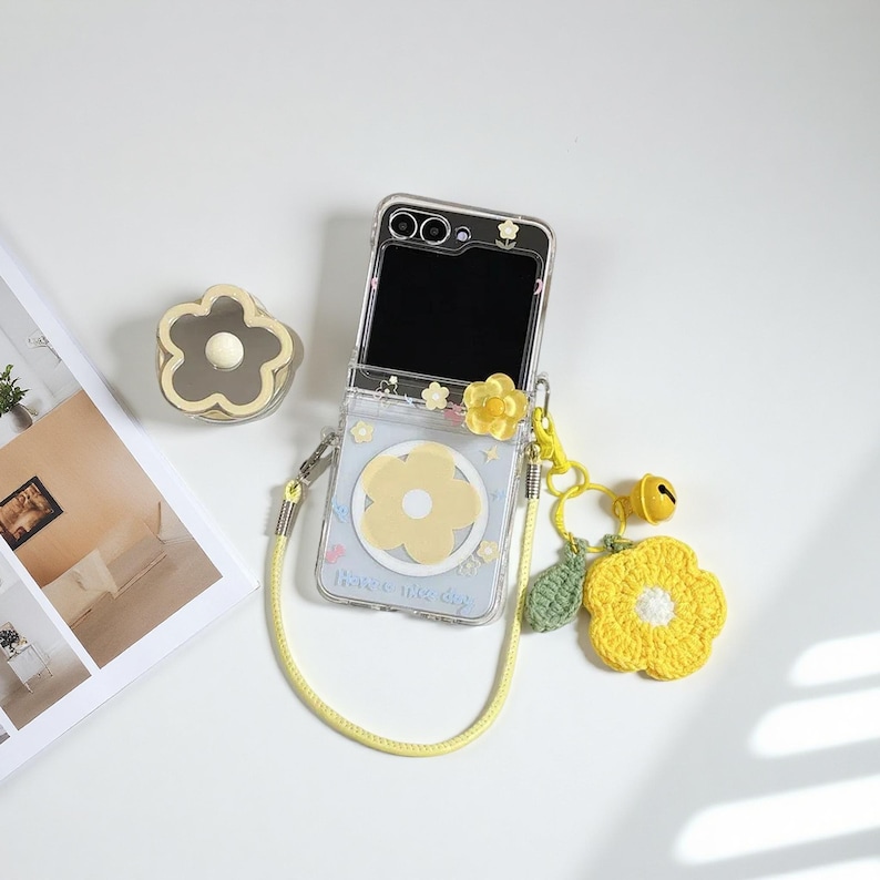 May include: Clear phone case with a yellow flower design and a yellow lanyard. The case has a yellow flower charm and a crocheted yellow flower charm with a green leaf. The phone case has the text "Have a nice day".