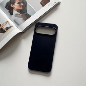 May include: A navy blue phone case with a rectangular camera cutout and a metallic trim. The case is lying on a white surface next to an open magazine with a woman's portrait.