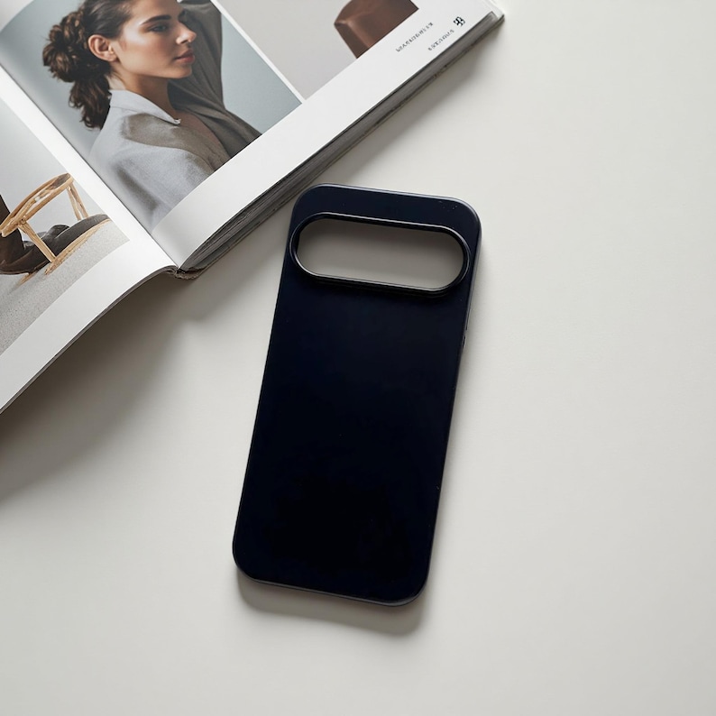 May include: A navy blue phone case with a rectangular camera cutout and a metallic trim. The case is lying on a white surface next to an open magazine with a woman's portrait.