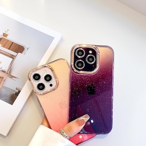 May include: Two clear phone cases with a gradient design. One case transitions from peach to pink, the other from gold to purple. Both cases have a gold-coloured camera ring and a clear Apple logo.