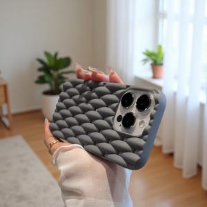 May include: A gray phone case with a textured, woven pattern. The case has a raised, bubble-like design and a camera cutout. The phone case is held up in front of a blurred background.