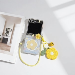 May include: A clear phone case with a yellow daisy design and a matching yellow strap. The case has a yellow flower charm and a yellow crocheted flower with a green leaf and a small bell. The text "Have a nice day" is visible.