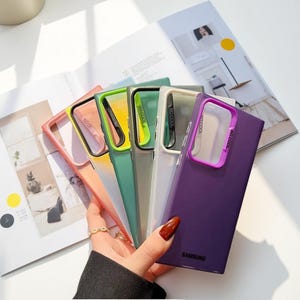 May include: A selection of vibrant phone cases in shades like pink, green, and purple. Each case features a clear central section and a coloured frame, with the text "SO COOL" printed on the side. The Samsung logo is visible on the purple case.